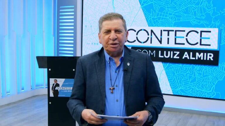 Luiz Almir de blazer azul apresenta o telejornal Acontece em frente ao telão com logotipo do programa na Band RN