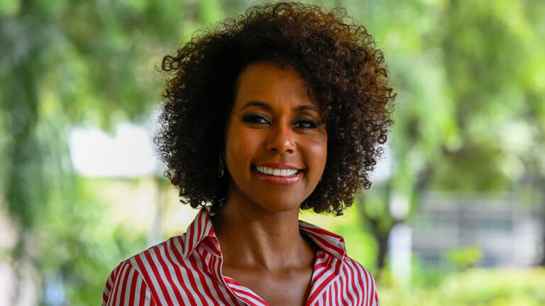 Maju Coutinho sorri usando camisa listrada vermelha e branca em ambiente externo com árvores ao fundo