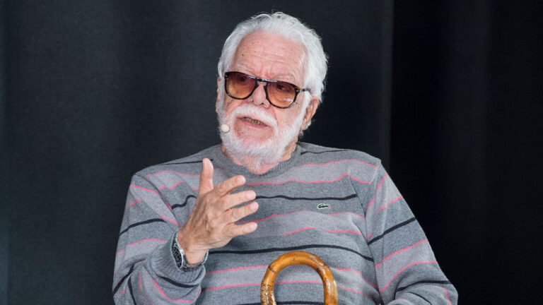 Manoel Carlos usa óculos escuros e suéter listrado cinza enquanto fala com microfone preso ao rosto segurando uma bengala