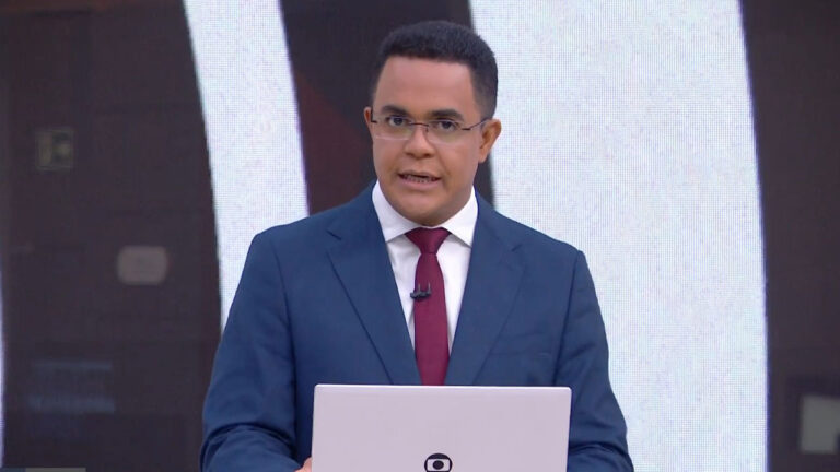 Marcelo Pereira apresenta telejornal da Globo usando terno azul e gravata vermelha diante de telão no estúdio