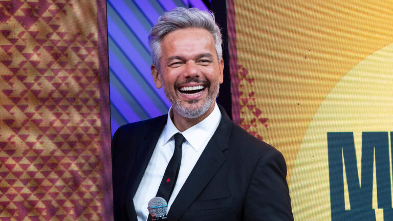 Otaviano Costa sorrindo de terno preto e gravata segurando microfone no palco do Melhor da Noite da Band