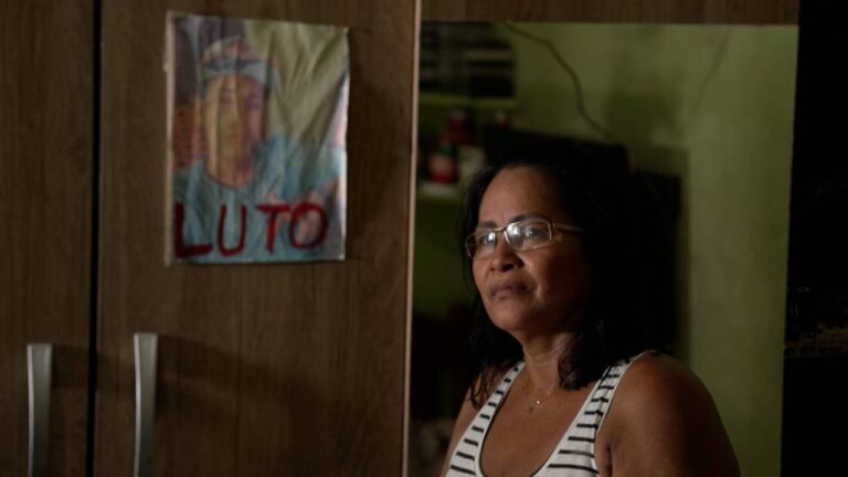 Mulher com expressão séria usa óculos e blusa listrada em frente a armário com cartaz escrito luto em reportagem sobre desaparecidos