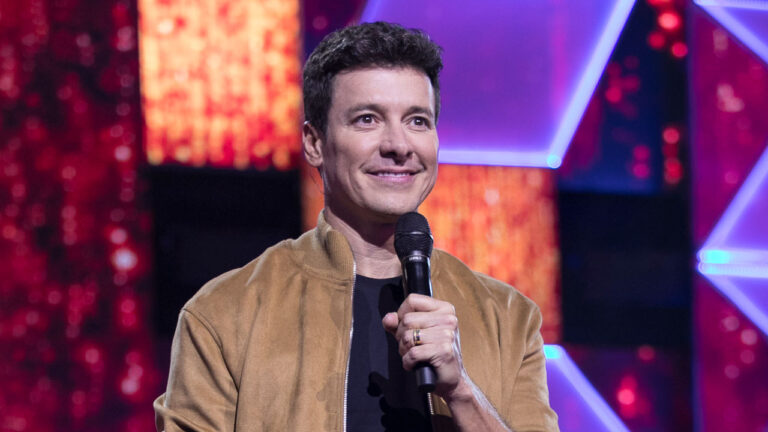 Rodrigo Faro com microfone na mão usa jaqueta bege no palco iluminado do Canta Comigo