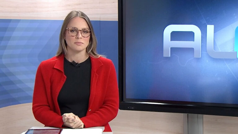 Sofia Sepreny com óculos e blazer vermelho apresenta o AL2 da TV Gazeta de Alagoas em estúdio com tela ao fundo