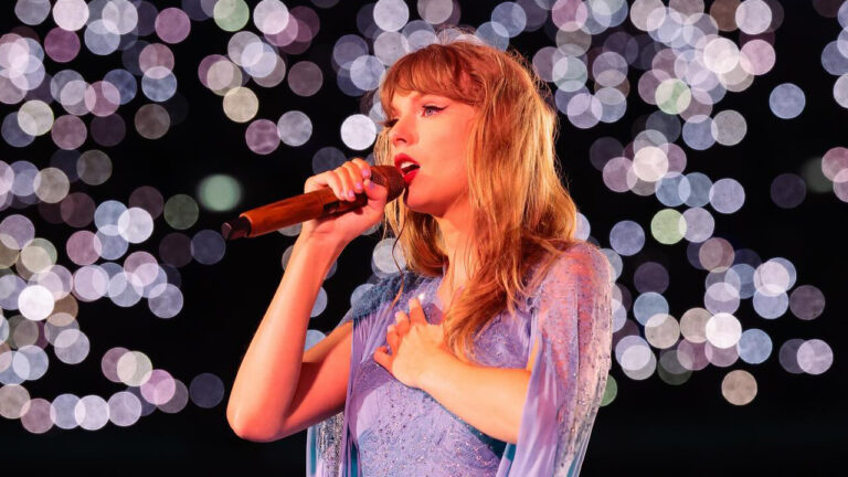 Taylor Swift canta no palco com microfone dourado na mão e vestido lilás diante de luzes desfocadas ao fundo