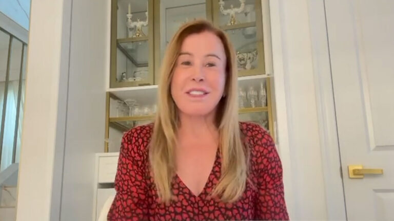 Zilu Camargo com blusa vermelha estampada sentada em ambiente interno com armário de vidro ao fundo