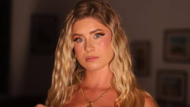 Débora Peixoto de cabelo loiro e olhos azuis em enquadramento fechado com maquiagem forte e colar dourado