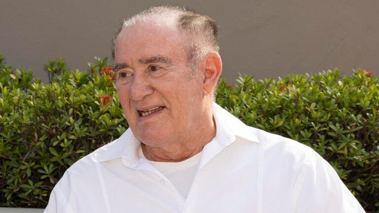 O humorista e ator **Renato Aragão** sorrindo de camisa branca em frente a uma sebe verde com flores vermelhas