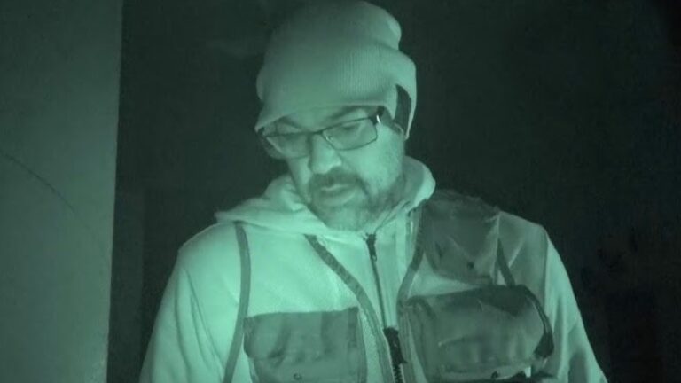 Aaron Goodwin em cena com iluminação de visão noturna esverdeada, usando gorro, óculos e colete tático, durante investigação paranormal em local escuro