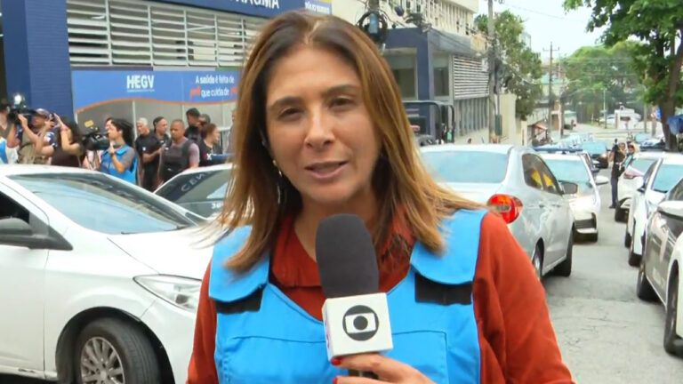 A repórter Adriana Rezende, da Globo Rio, faz uma entrada ao vivo com colete azul e microfone em uma rua movimentada