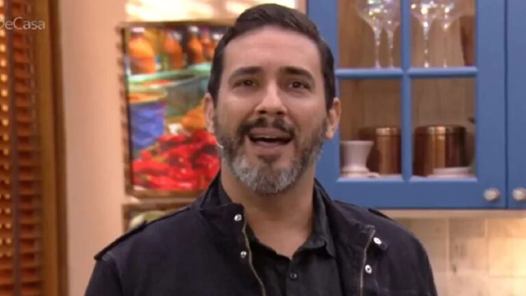 André Marques em estúdio de programa culinário, vestindo jaqueta preta e camisa escura, com prateleiras e utensílios de cozinha ao fundo
