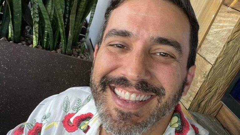 André Marques sorrindo em uma foto com camisa estampada de flores, ao ar livre, com plantas ao fundo