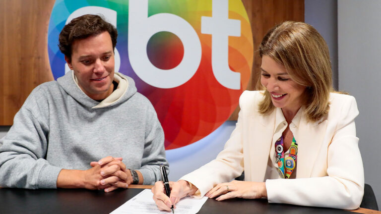 André Valadão, de moletom cinza, observa Daniela Beyruti, de blazer claro, assinar contrato em uma sala de reunião na sede do SBT