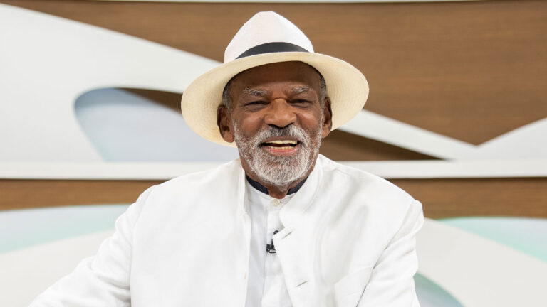 O ator Antonio Pitanga sorri e usa um terno e um chapéu fedora brancos durante participação no programa Roda Viva da TV Cultura