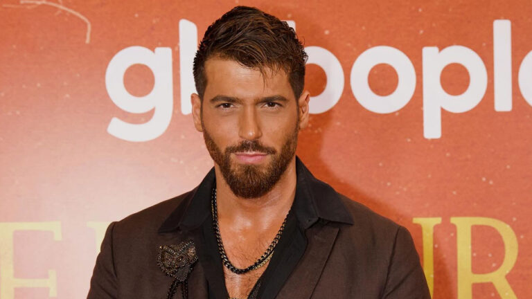 O ator Can Yaman com barba e cabelo penteado para trás posa em evento com painel laranja e o logo do Globoplay desfocado ao fundo