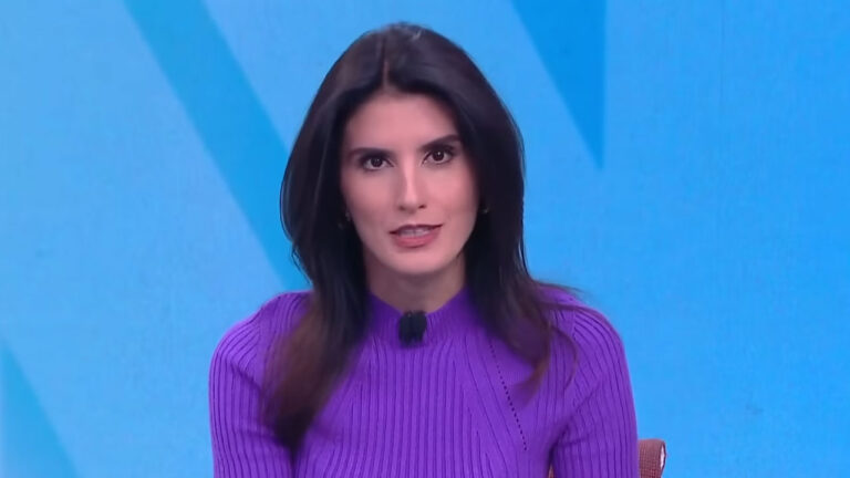 A apresentadora Carol Nogueira, com blusa roxa de tricô e cabelo escuro, apresenta o telejornal Novo Dia no estúdio da CNN Brasil