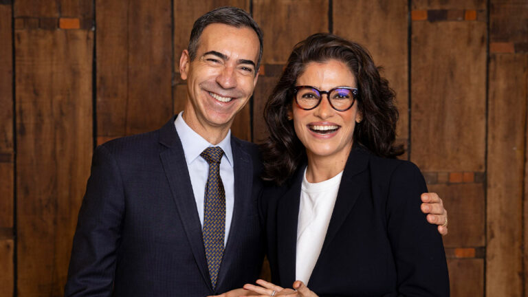 César Tralli e Renata Vasconcellos sorrindo juntos diante de parede de madeira em foto de ambiente fechado