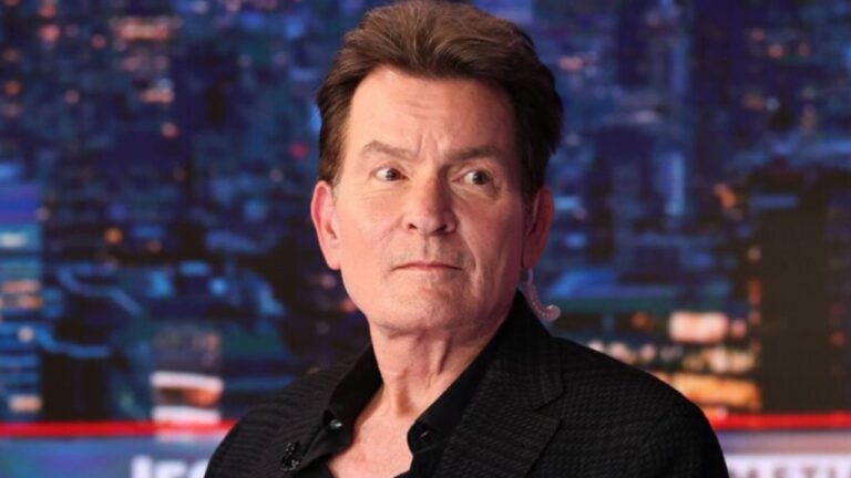 Charlie Sheen de paletó preto e em um estúdio iluminado com fundo desfocado de cidade à noite
