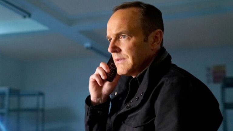 Clark Gregg veste uma jaqueta escura e fala ao celular em um ambiente fechado e iluminado por luz azulada, aparentando estar concentrado na conversa