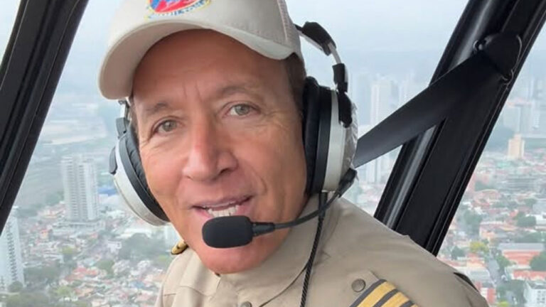 Comandante Hamilton usando fone de ouvido com microfone, boné claro e uniforme bege com insígnias de piloto dentro de helicóptero sobrevoando área urbana