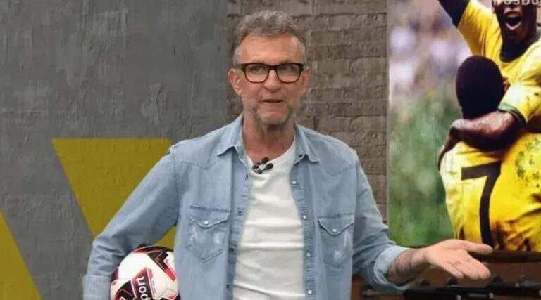 Craque Neto com barba grisalha segura uma bola de futebol em um estúdio esportivo, vestindo camiseta branca e camisa jeans clara, com uma imagem de jogadores de futebol ao fundo