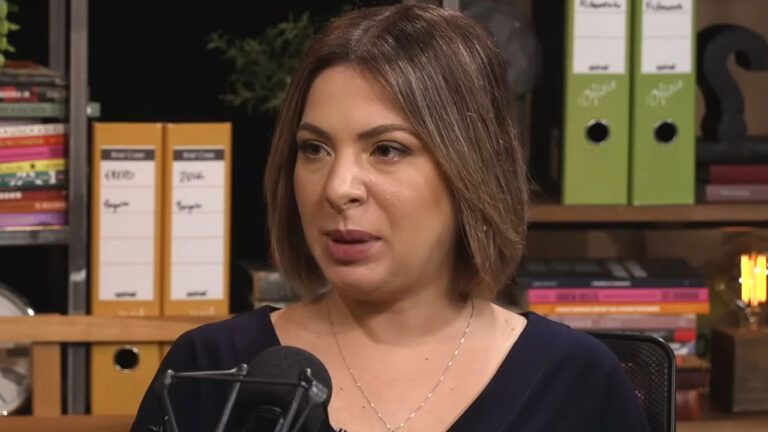 A jornalista Daniela Lima com cabelo curto e blusa escura fala em um microfone em um estúdio com estantes de livros e fichários ao fundo