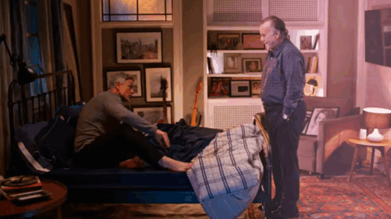 Cena da novela Dona de Mim mostra dois homens em um quarto aconchegante — um sentado na cama, o outro em pé — em meio a uma decoração com quadros, livros e luz ambiente amarelada