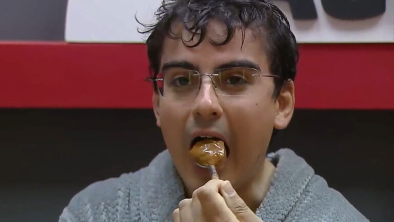 Dudu Camargo usando óculos e roupão cinza enquanto come doce de leite com uma colher dentro da casa de A Fazenda