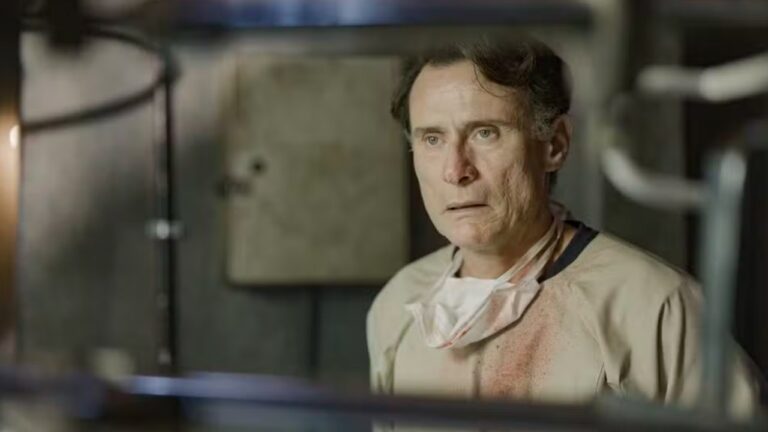 Homem com expressão assustada usa roupa bege manchada e máscara pendurada no pescoço em ambiente escuro e metálico em cena da série Reencarne da Globoplay