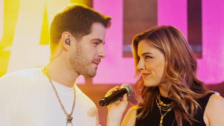 Enzzo Gabriel (Maurício Destri) e Raíssa (Alice Wegmann) se olham apaixonados no palco com luzes rosas ao fundo
