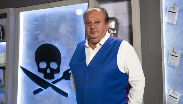 Erick Jacquin em estúdio de programa culinário, vestindo colete azul sobre camisa branca, posando diante de um painel com símbolo de caveira formada por talheres cruzados