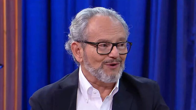 O jornalista Ernesto Paglia, com camisa branca e terno preto, fala durante participação em programa com cenário de cortina azul