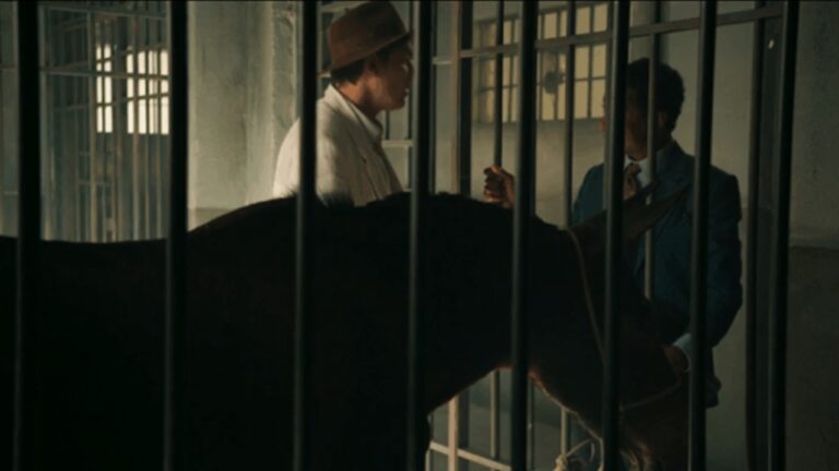Cena da novela Êta Mundo Melhor! mostra dois homens conversando próximos a um cavalo dentro de um estábulo, separados por grades de ferro, em ambiente com luz natural suave