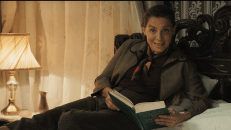 Cena da novela Êta Mundo Melhor! mostra uma mulher deitada na cama, sorrindo enquanto segura um livro aberto em ambiente com iluminação amarelada e decoração de época.