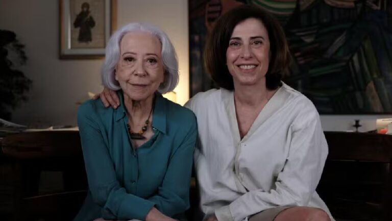 Fernanda Montenegro e Fernanda Torres sentadas lado a lado, sorrindo para a câmera em um ambiente interno. A mulher à esquerda tem cabelos grisalhos e usa uma blusa azul; a da direita tem cabelos castanhos curtos e veste uma camisa clara