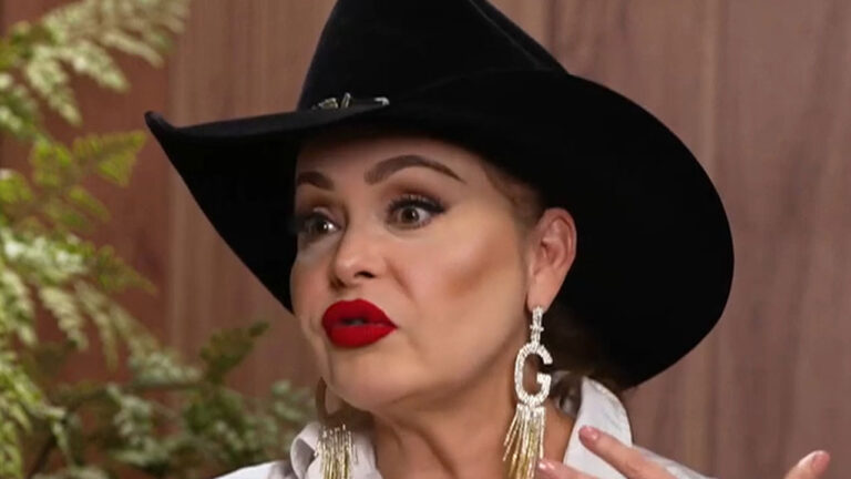 Gaby Spanic usando chapéu preto, brincos longos dourados com a letra G e batom vermelho enquanto fala gesticulando