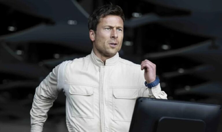Glen Powell em cena vestindo macacão de piloto claro, com expressão concentrada, diante de um fundo escuro que sugere ambiente aeronáutico ou de treinamento