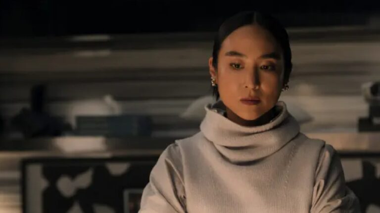 Greta Lee em cena de The Morning Show, usando suéter claro em ambiente com iluminação suave e tom sóbrio