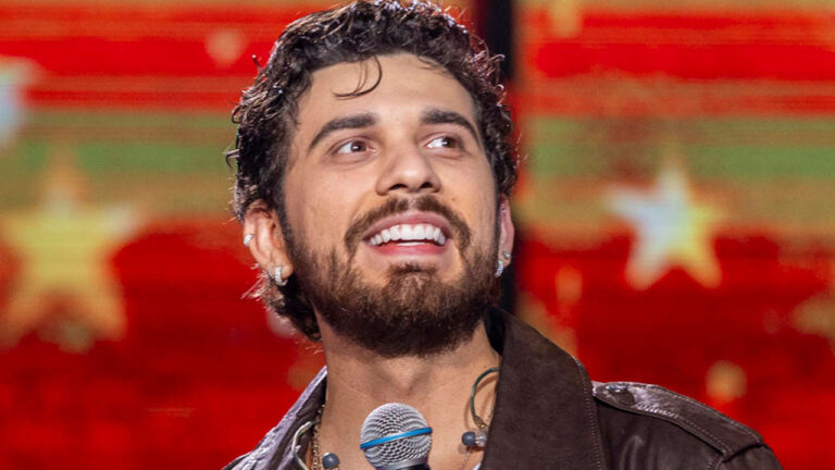 O cantor Gustavo Mioto sorrindo de cabelo cacheado e jaqueta de couro com microfone contra fundo vermelho estrelado