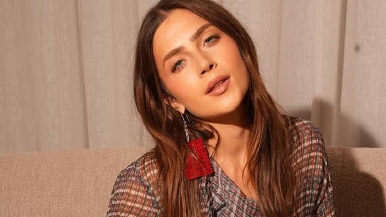 Jade Picon posa sentada em ambiente interno, usando blusa estampada e brinco vermelho longo, com expressão suave e iluminação quente