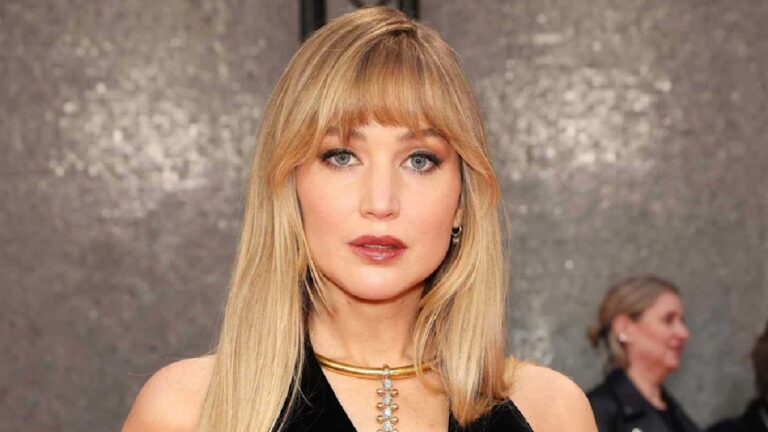 Mulher de cabelos loiros e franja usa vestido preto e colar dourado com pingente em evento com fundo prateado brilhante e pessoas ao fundo desfocadas