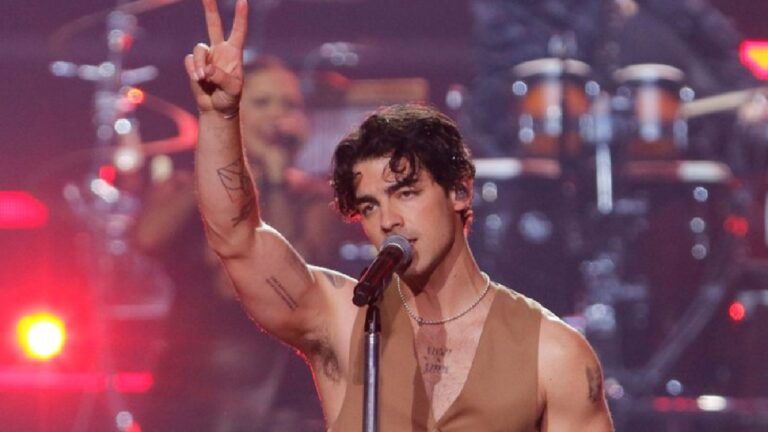 Joe Jonas se apresentando no palco, cantando ao microfone com expressão intensa e fazendo o gesto de paz com a mão levantada, sob luzes vermelhas e cenário de show ao fundo