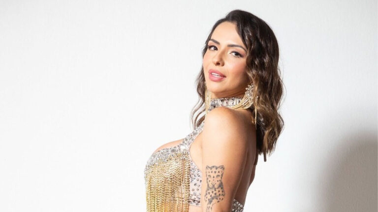 Karol Rosalin posa de lado com top bordado e franjas douradas, olhando por cima do ombro para a câmera, com tatuagem de onça no braço