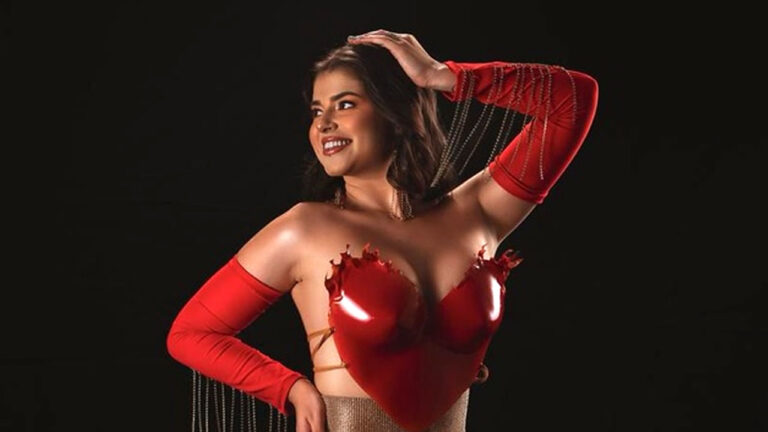 Kerolay Chaves sorri com a mão na cabeça usando top vermelho em formato de coração e mangas vermelhas com correntes em fundo preto