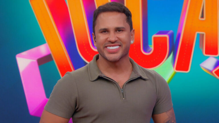 O influenciador Lucas Guimarães sorri com camisa polo verde com zíper em frente a um cenário de programa de TV colorido e iluminado