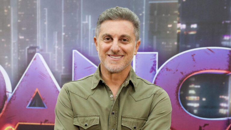 Luciano Huck sorrindo no cenário do Domingão com Huck com painel colorido e luzes de estúdio ao fundo