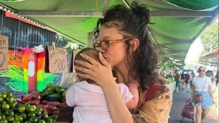 Manu Gavassi em uma feira livre beijando o bebê que segura nos braços, cercada por barracas de frutas e pessoas ao fundo sob tendas verdes