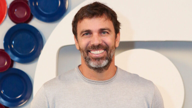 O ator Marcelo Faria com barba grisalha e camiseta cinza sorri para a câmera em frente a um painel decorado com pratos azuis e vermelhos