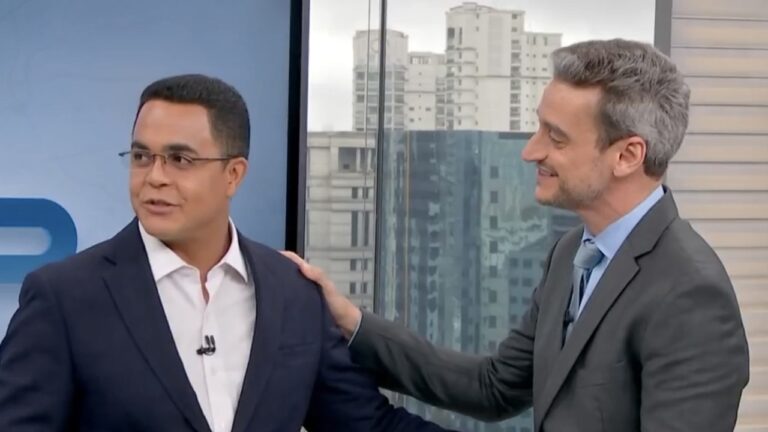 Marcelo Pereira e Tiago Scheuer usam ternos e sorriem durante conversa no estúdio do telejornal Bom Dia São Paulo com vista de prédios ao fundo
