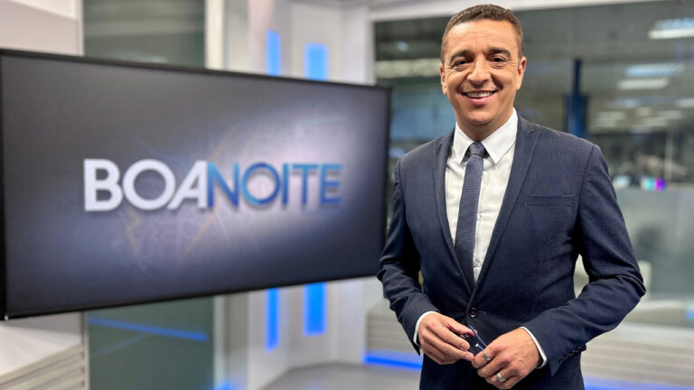 Marcelo Rocha de terno escuro sorri no estúdio do Boa Noite Paraná da RPC com o logo do programa na tela